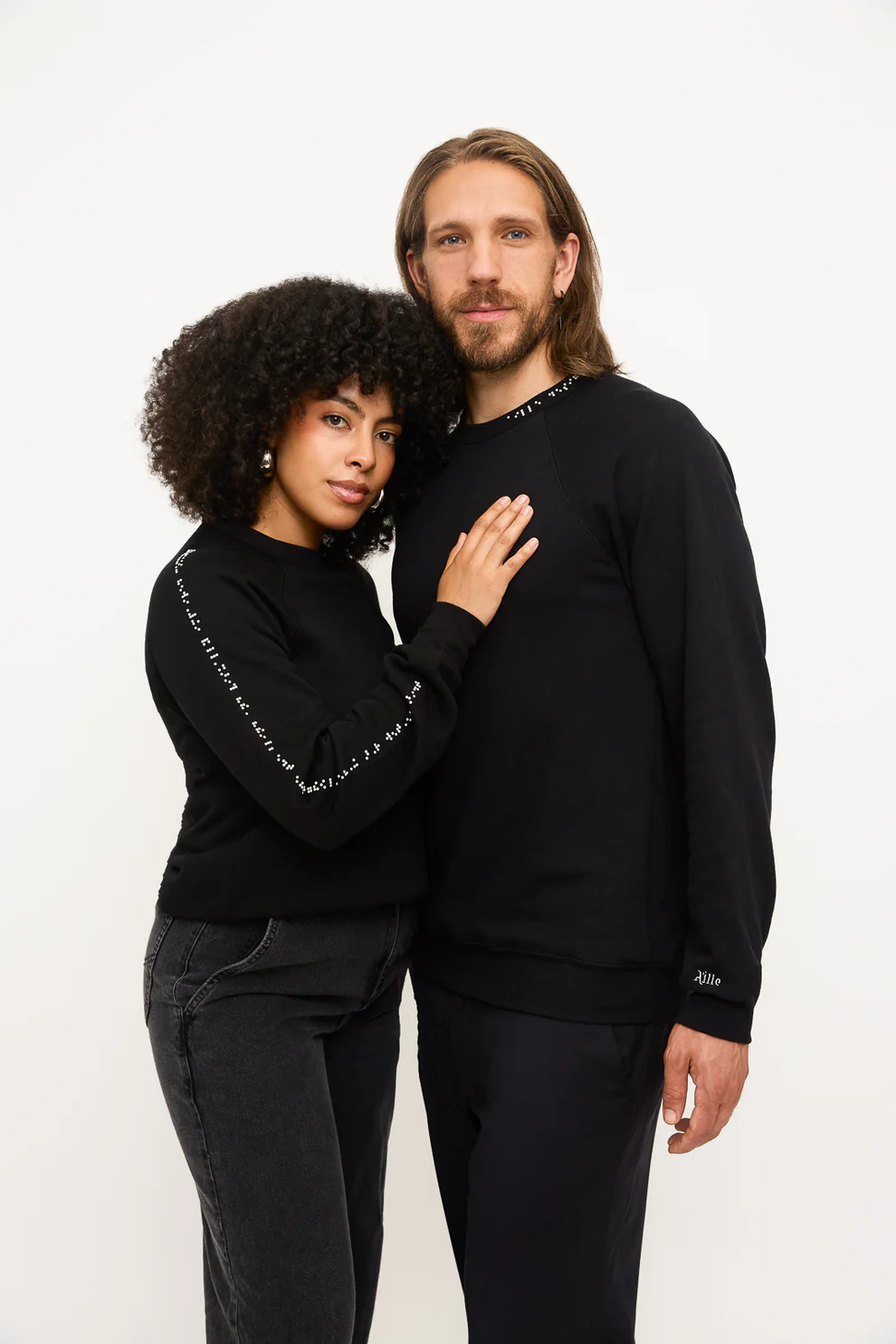 Man and woman wear black sweatshirts with braille in white on the sleeve for the woman and at the collar for the man